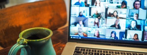 A laptop displaying a grid view of a virtual meeting with multiple participants on screen, alongside a turquoise ceramic mug on a wooden table.